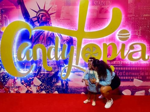 A woman kneels beside a smiling child in front of a colorful "CandyTopia" backdrop featuring the Statue of Liberty.