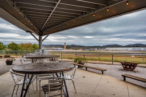 View from a patio with tables overlooking an airport runway and distant mountains under a cloudy sky.