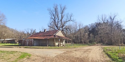 A rustic house with a metal roof sits at a dirt road intersection, surrounded by bare trees and open fields.