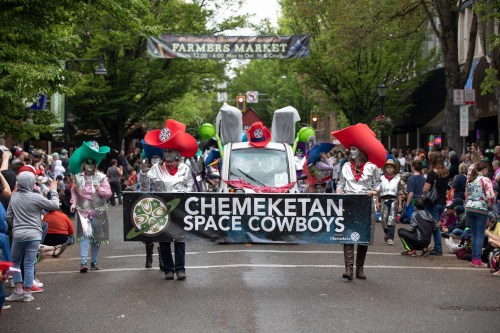 A parade scene featuring the Chemeketan Space Cowboys with colorful costumes and a banner, surrounded by a cheering crowd.