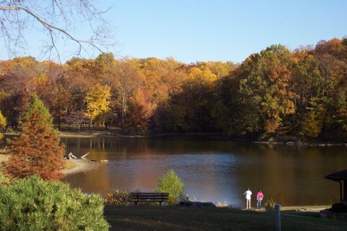 Two people walk by a serene lake surrounded by vibrant autumn foliage.
