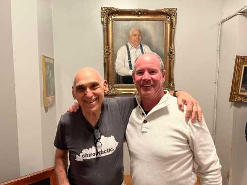 Two smiling men pose together in front of a framed portrait of an older man on the wall.