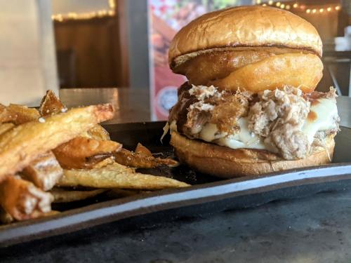 A close-up of a pulled pork sandwich with crispy onion rings, served with golden fries on a black plate.