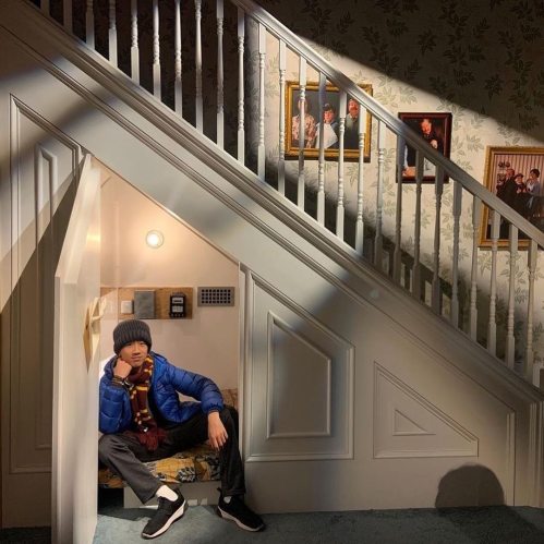 A person sits under a staircase, wearing a blue jacket and beanie, with framed photos on the wall above.