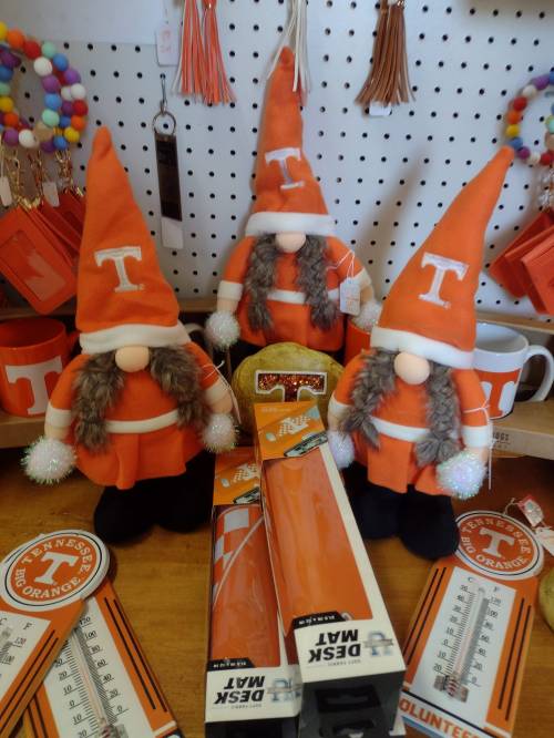 Three orange-clad gnome figurines with "T" hats stand among various Tennessee-themed merchandise on a wooden table.