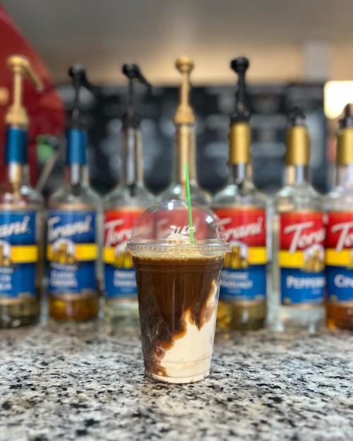 A layered iced coffee drink in a clear cup, with syrup and cream, set against a backdrop of flavored syrup bottles.