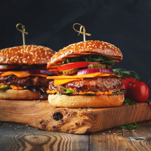 Two delicious burgers with sesame seed buns, stacked with cheese, lettuce, onions, and pickles on a wooden board.