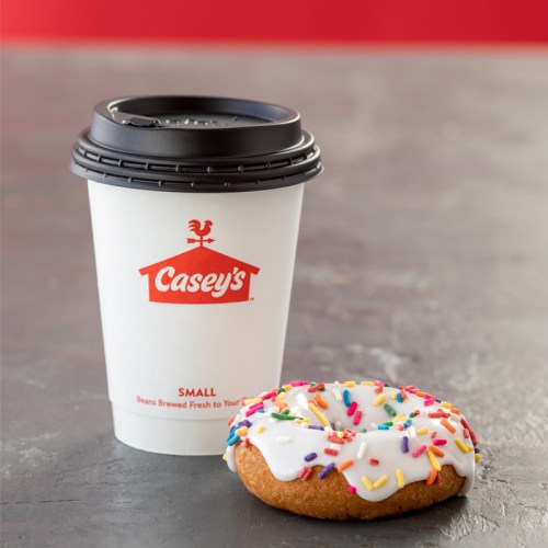 A coffee cup from Casey's next to a frosted donut topped with colorful sprinkles on a gray surface.