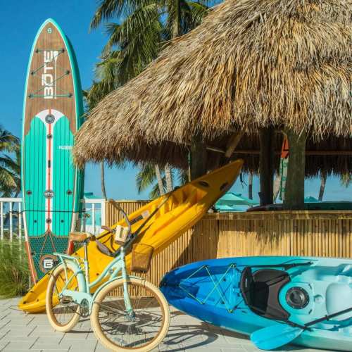 A colorful paddleboard and kayaks are propped against a thatched hut, with a bicycle nearby, set in a tropical beach scene.