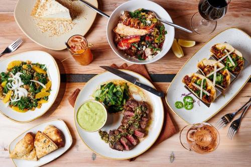 A spread of various dishes including steak, tacos, salads, and dessert on a wooden table with drinks.