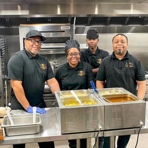 Four people in a kitchen, smiling and standing behind food trays, wearing matching black shirts and hats.