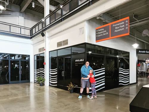A man and a child stand in front of a glass storefront labeled "AVAILABLE" in a modern indoor space.