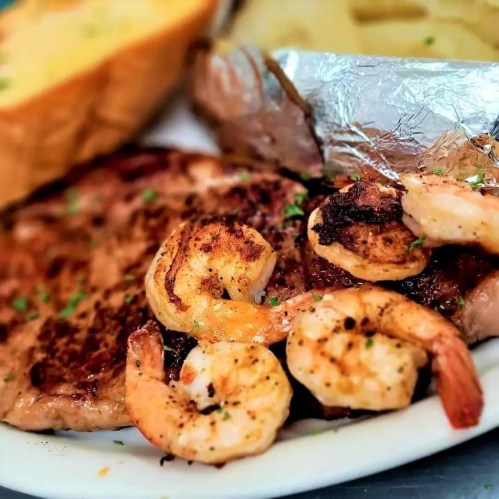A plate featuring grilled shrimp, a steak, and a side of pasta, garnished with herbs and served with bread.