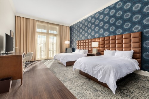 A modern hotel room featuring two beds, a large window with curtains, and stylish decor on the walls.