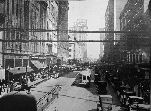 Historic black-and-white street scene with vintage cars, streetcars, and pedestrians in a bustling city.
