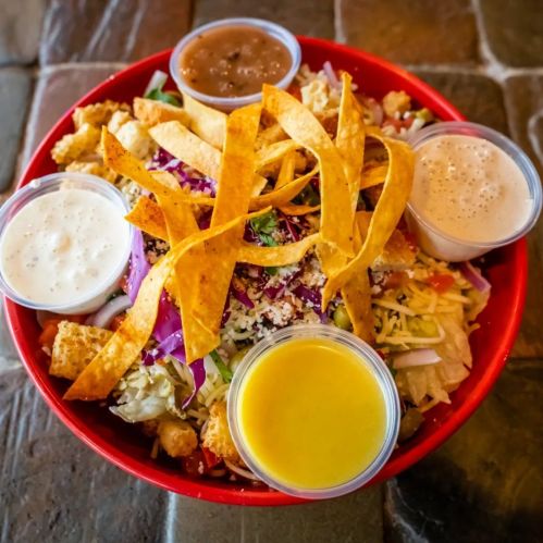 A colorful salad bowl topped with tortilla strips, cheese, and four small containers of dressing.