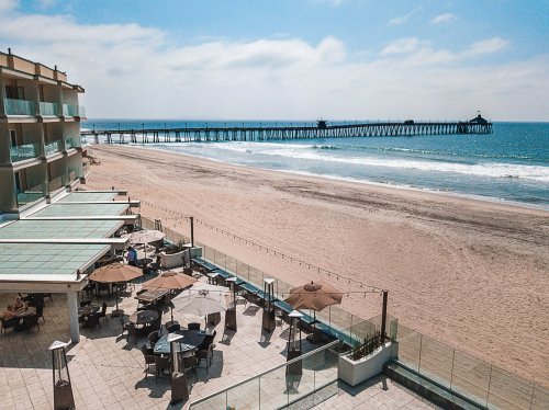 A beachfront view featuring a pier, sandy shore, and outdoor seating area with umbrellas under a clear blue sky.