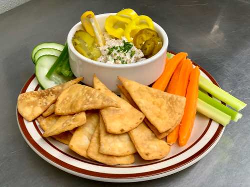 A plate of crispy tortilla chips with a bowl of dip, surrounded by cucumber, celery, carrot sticks, and pickled peppers.