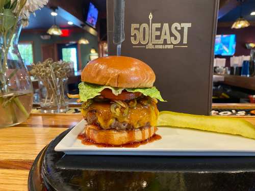 A juicy burger with cheese, lettuce, tomato, and jalapeños on a plate, accompanied by a pickle, set in a restaurant.