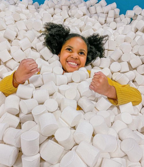 A smiling girl with curly hair playfully emerges from a pile of white marshmallows, showing excitement and joy.