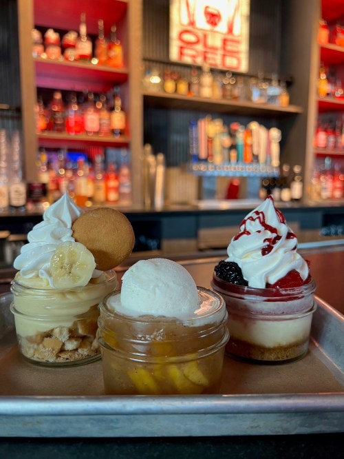 Three dessert jars on a tray: banana pudding, peach cobbler, and strawberry cheesecake, with a bar backdrop.