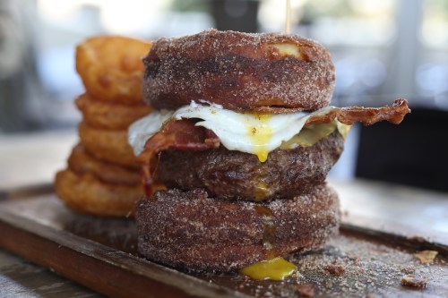 A stacked burger with a donut bun, bacon, fried egg, and a side of onion rings on a wooden platter.
