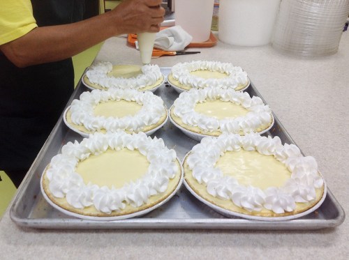 A person decorates six cream pies with whipped cream on a baking tray in a kitchen setting.