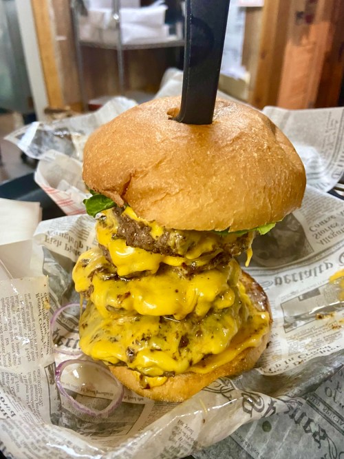A towering cheeseburger with three beef patties, melted cheese, lettuce, and a knife inserted on top, served in paper.