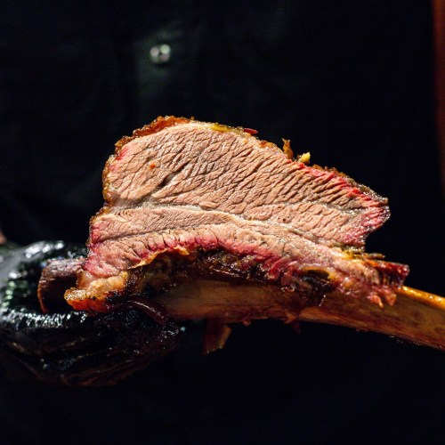 A close-up of a juicy, smoked beef brisket slice held by a person wearing black gloves.