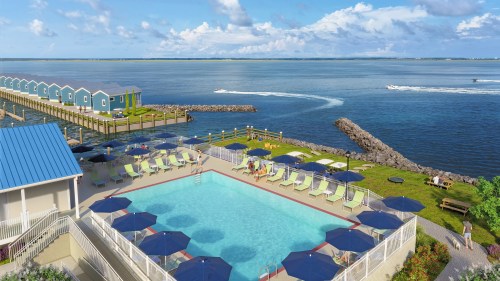 A scenic waterfront view featuring a pool, lounge chairs, and colorful cabins along the shore under a blue sky.