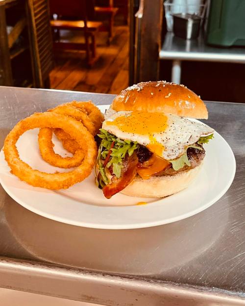 A delicious burger with lettuce, bacon, and a fried egg, served with crispy onion rings on a white plate.