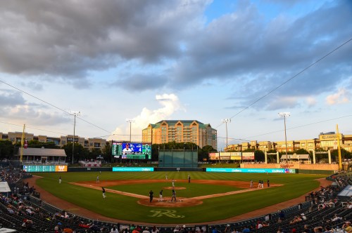 A baseball field at sunset, with players on the field and spectators in the stands, surrounded by buildings and clouds.