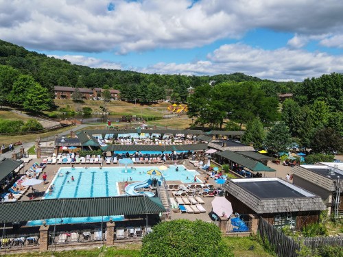 Aerial view of a busy pool area with sun loungers, surrounded by greenery and recreational facilities on a sunny day.