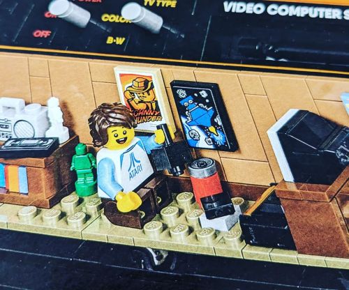 A LEGO figure with brown hair sits in a retro room, surrounded by gaming memorabilia and colorful decorations.