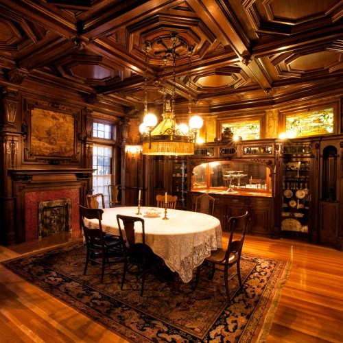 A richly decorated dining room with a wooden table, chairs, ornate ceiling, and a vintage chandelier.