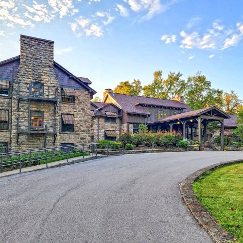 A stone mansion with a sloped driveway, surrounded by trees and a clear blue sky.