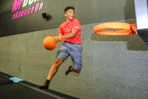 A young boy in a red shirt jumps to dunk a basketball at a trampoline park.