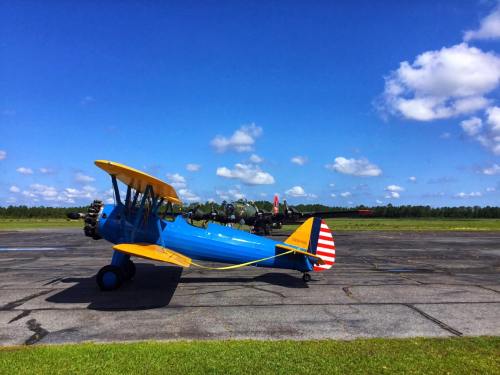 A blue biplane with a yellow wing parked on an airstrip under a bright blue sky with fluffy clouds.