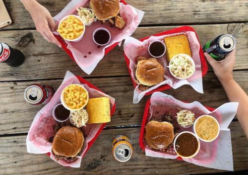 Four trays of barbecue meals with sandwiches, sides of macaroni and coleslaw, and drinks on a wooden table.