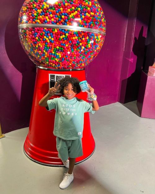 A young child smiles and poses next to a large gumball machine filled with colorful gumballs.