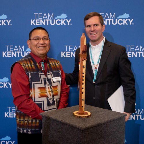 Two men pose together in front of a blue "Team Kentucky" backdrop, with a wooden flute displayed on a pedestal between them.