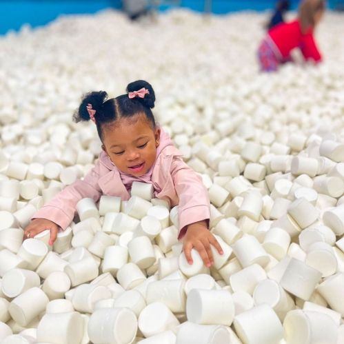 A young girl in a pink jacket plays in a sea of white cups, reaching out with a joyful expression.