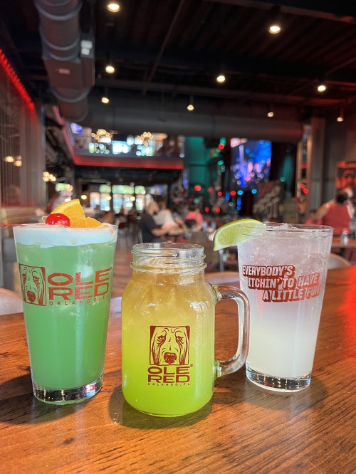 Three colorful cocktails on a wooden table, with a lively bar scene in the background.