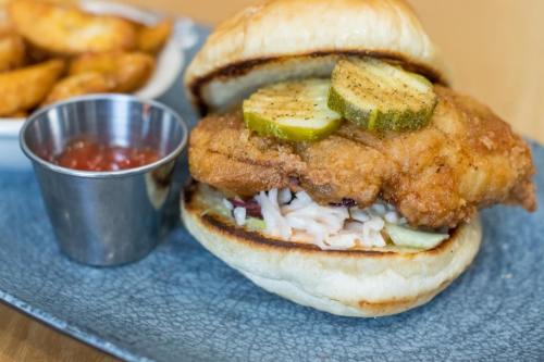A crispy fried chicken sandwich with pickles and coleslaw, served with a side of potato wedges and dipping sauce.