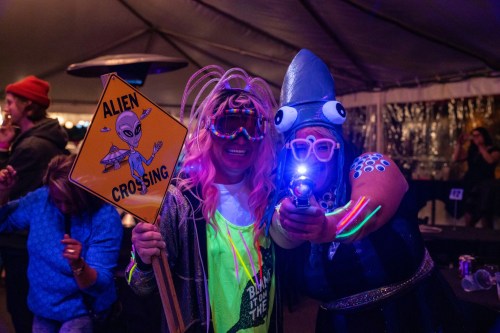 Two people in colorful costumes pose under a tent, one holding an "Alien Crossing" sign and the other with a toy gun.