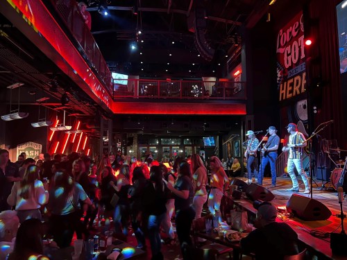 A lively crowd dances in a vibrant venue with a band performing on stage, illuminated by colorful lights.