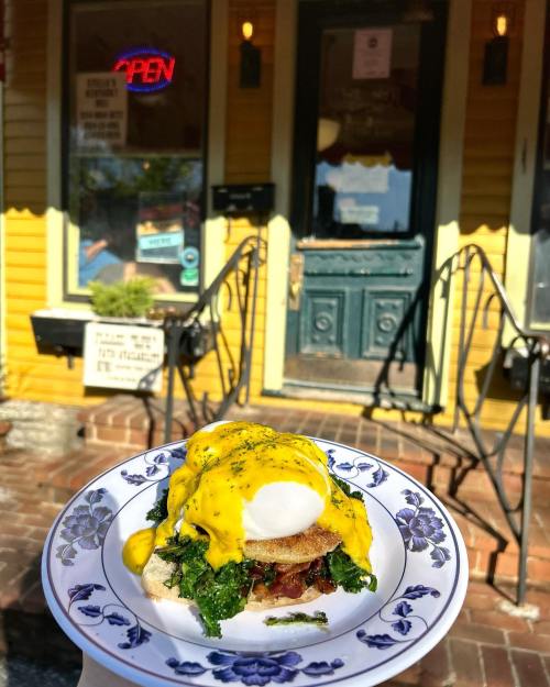 A plate of eggs Benedict with spinach and hollandaise sauce in front of a charming yellow restaurant with an "Open" sign.