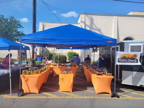 A vibrant market scene with blue tents and orange tables displaying jars of goods under a clear sky.