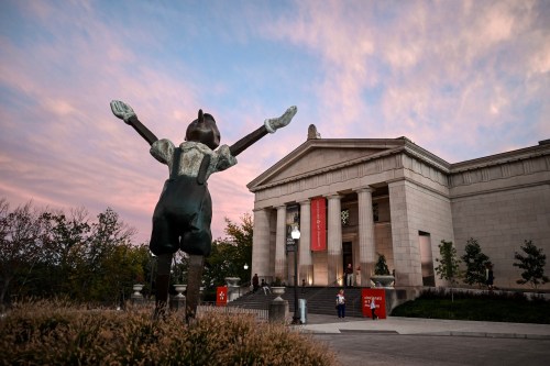A statue of a joyful figure stands in front of a museum at sunset, with colorful clouds in the sky.