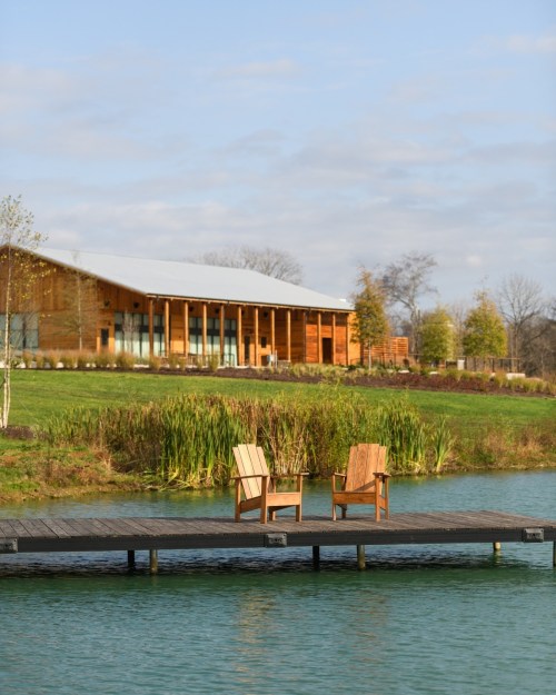 Two wooden chairs sit on a dock by a calm lake, with a modern building and greenery in the background.